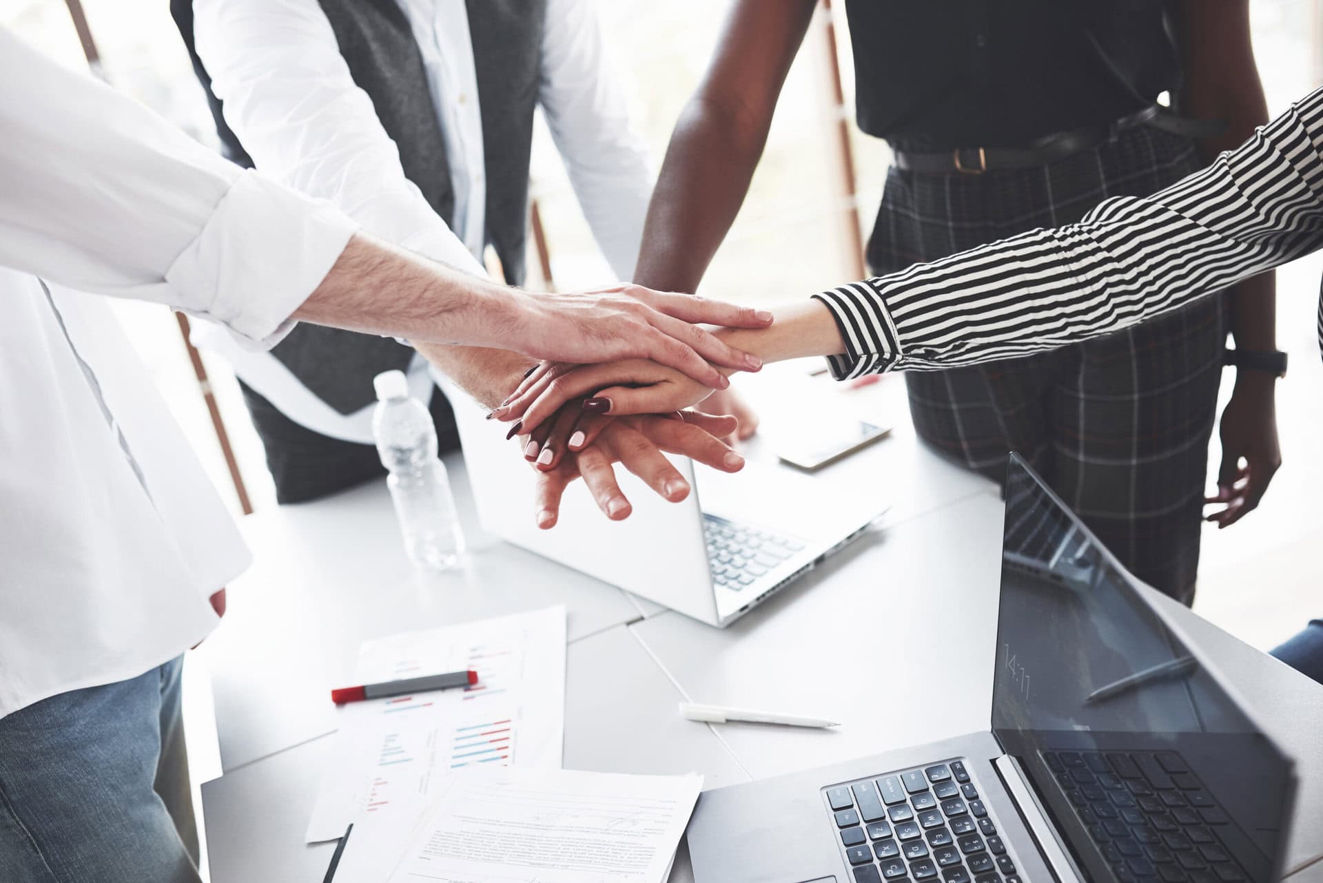 Photo of people with their hands on top of each other's. Laptop's and a water bottle are in the back.