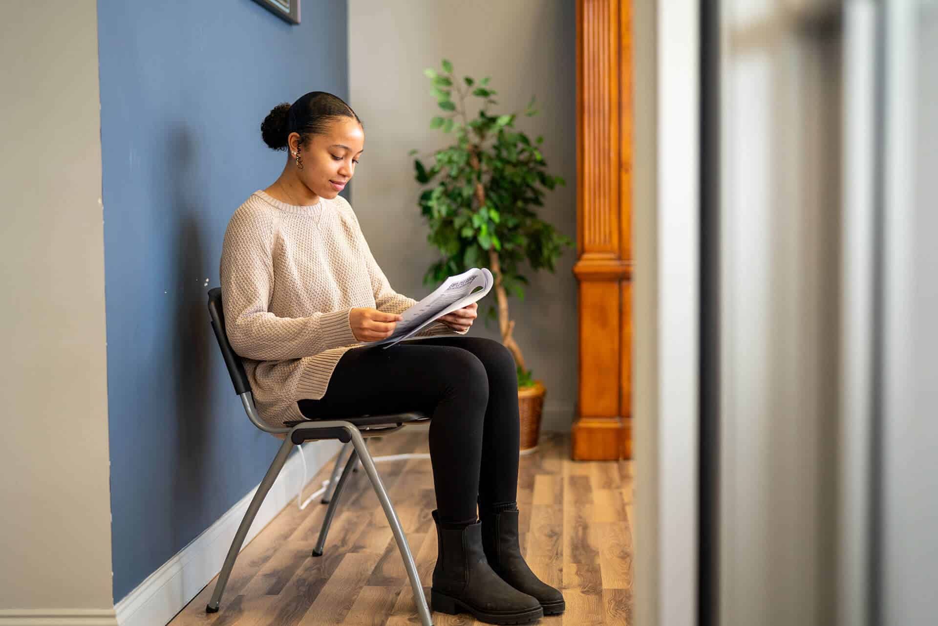 Photo of a woman reading a piece of paper while sitting on a chair at First Choice Staffing. With a green plant behind her.