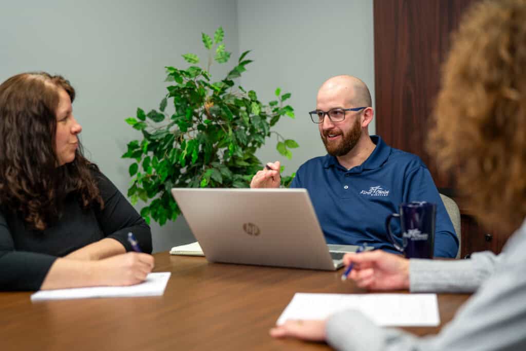 Man talking with laptop open on conference table, can see left side someone is writing and wearing a long sleeved black shirt and across the table is someone writing on a paper with long grey sleeves