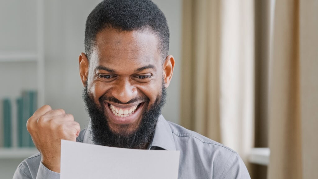 man with black hair and beard smiling while looking at a white piece of paper and giving a fist pump. He appears excited