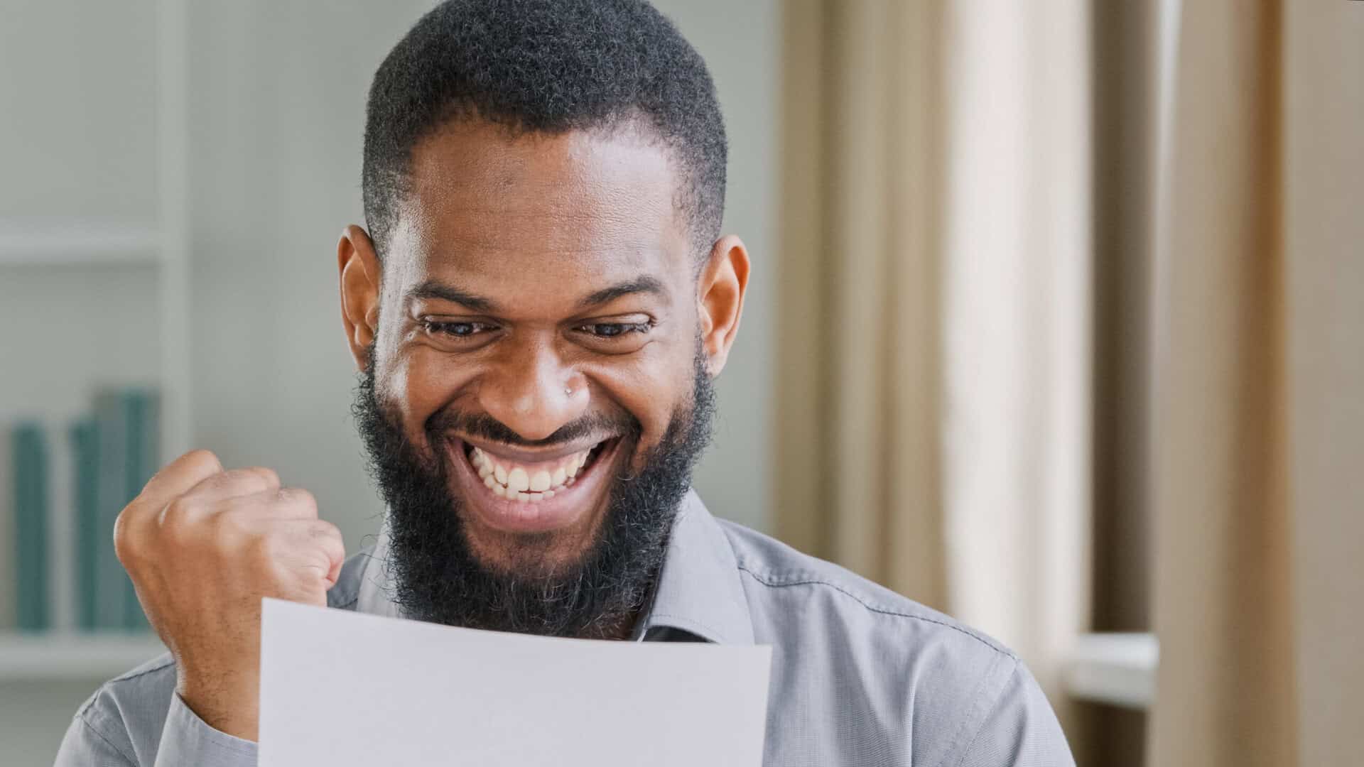 man with black hair and beard smiling while looking at a white piece of paper and giving a fist pump. He appears excited