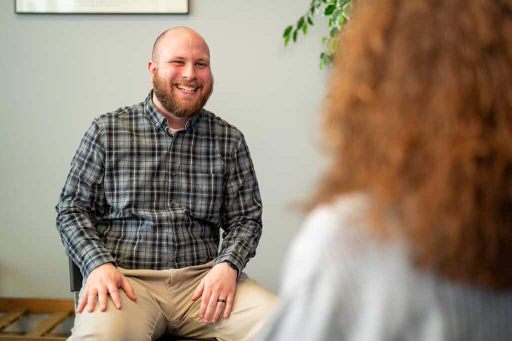 bearded man facing camera smiling, relaxed hands on lap, wearing a plaid long sleeved shirt. Foreground has a red haired woman facing away from camera