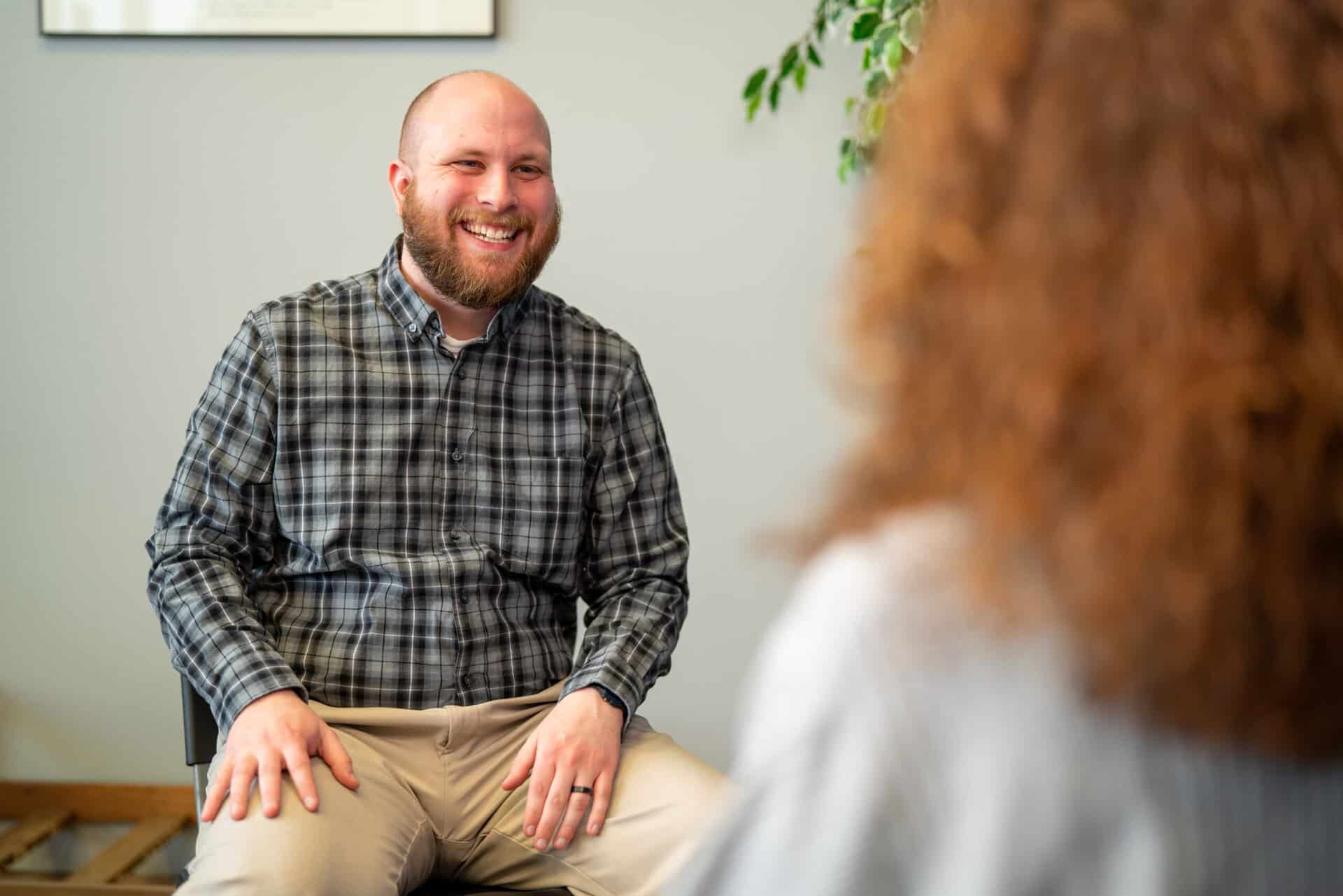 bearded man facing camera smiling, relaxed hands on lap, wearing a plaid long sleeved shirt. Foreground has a red haired woman facing away from camera