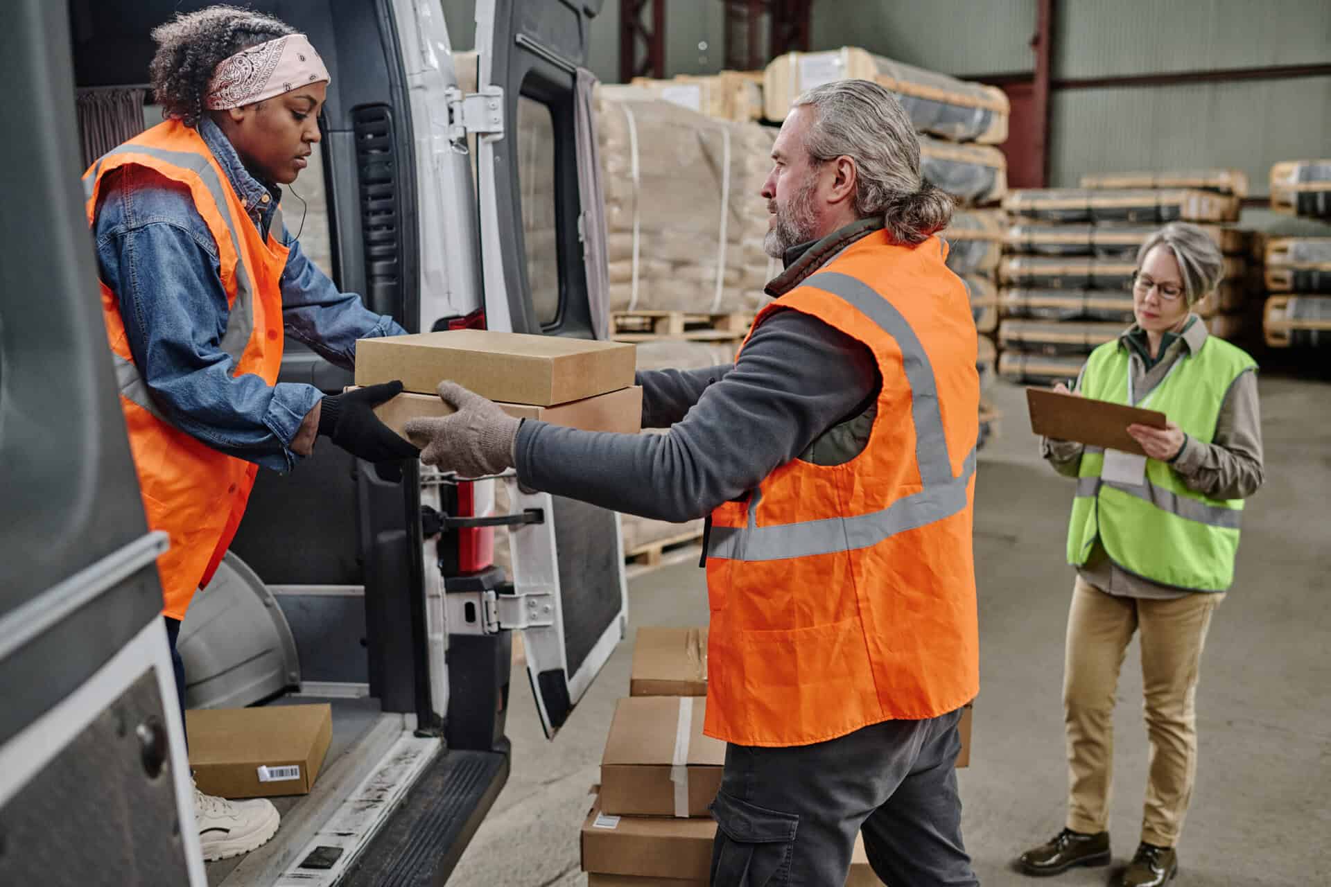 Two workers loading boxes from the back of a truck inside a warehouse. Person in the background with a clipboard.
