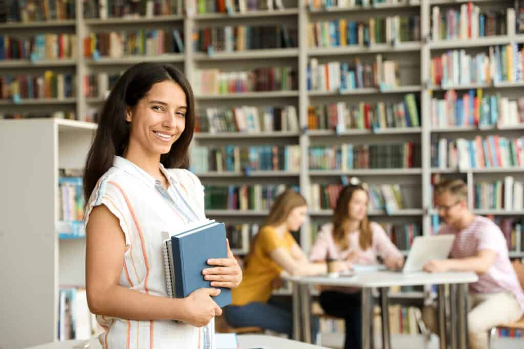 Happy young woman with books in library. blurry book shelves in the background. people sitting around a round table talking