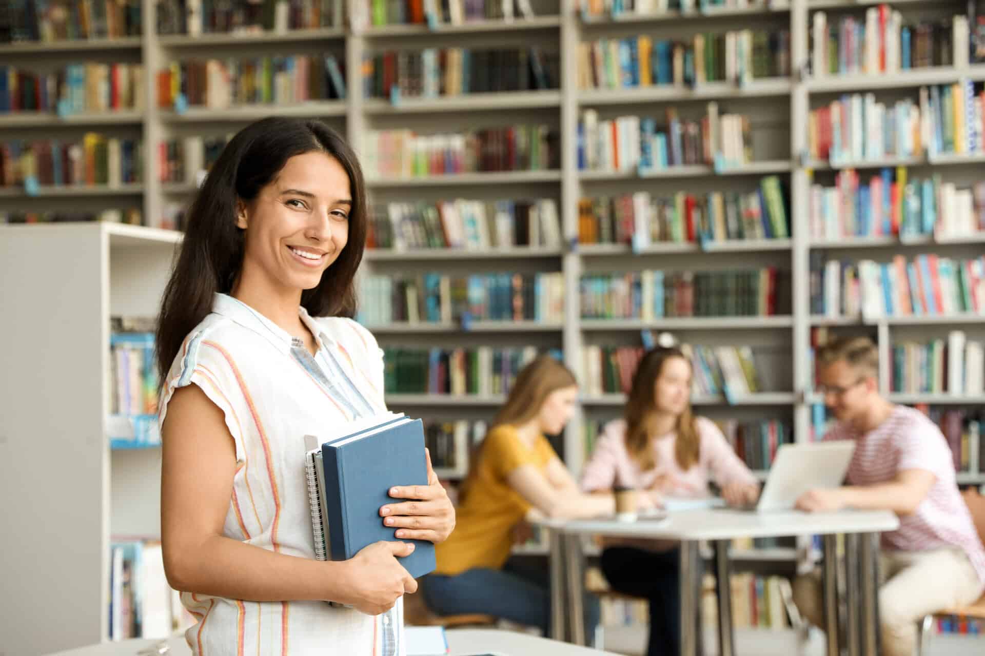 Happy young woman with books in library. blurry book shelves in the background. people sitting around a round table talking